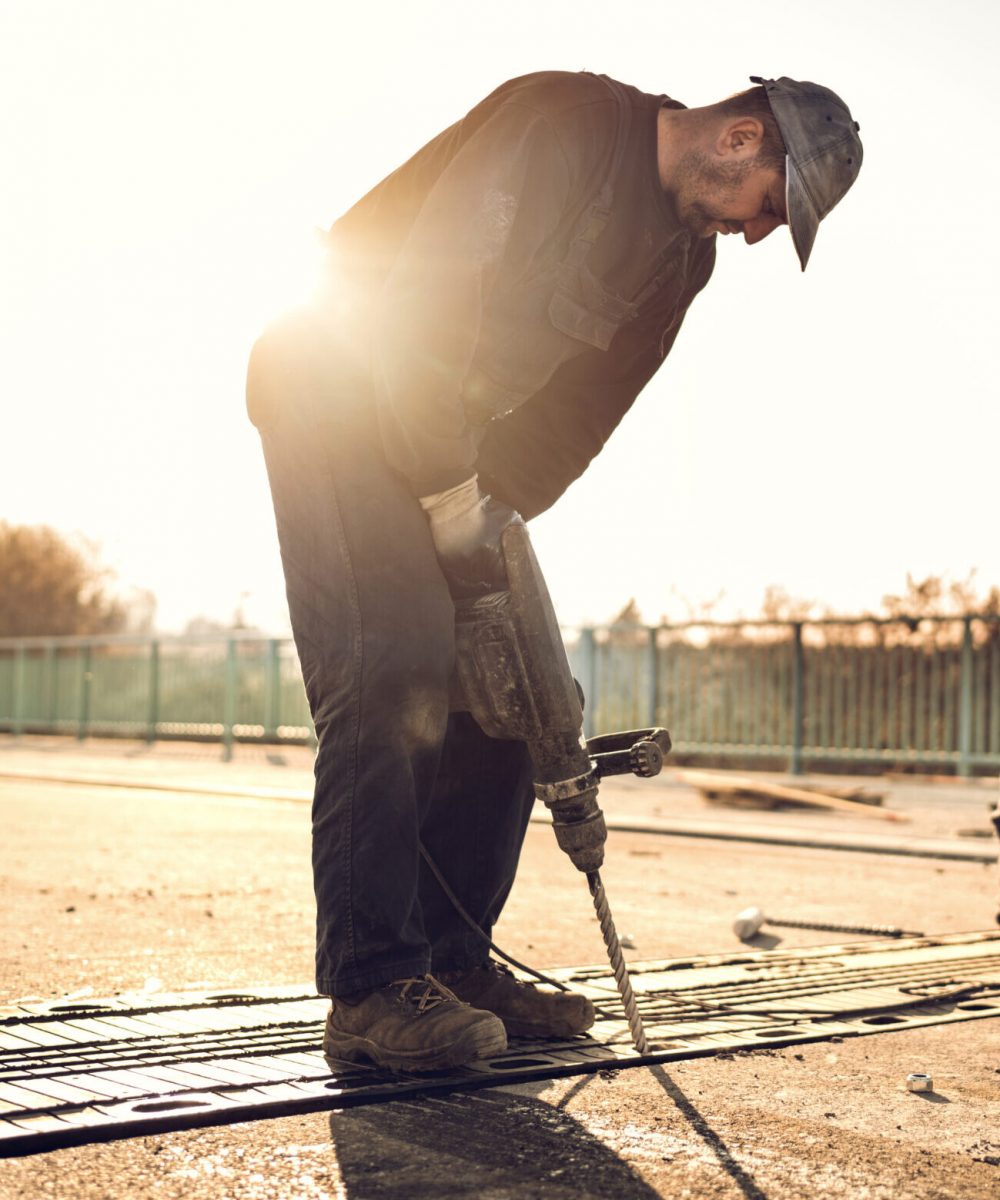 Two manual workers repairing road construction on the bridge at sunset. Focus is on construction worker using a drill.
