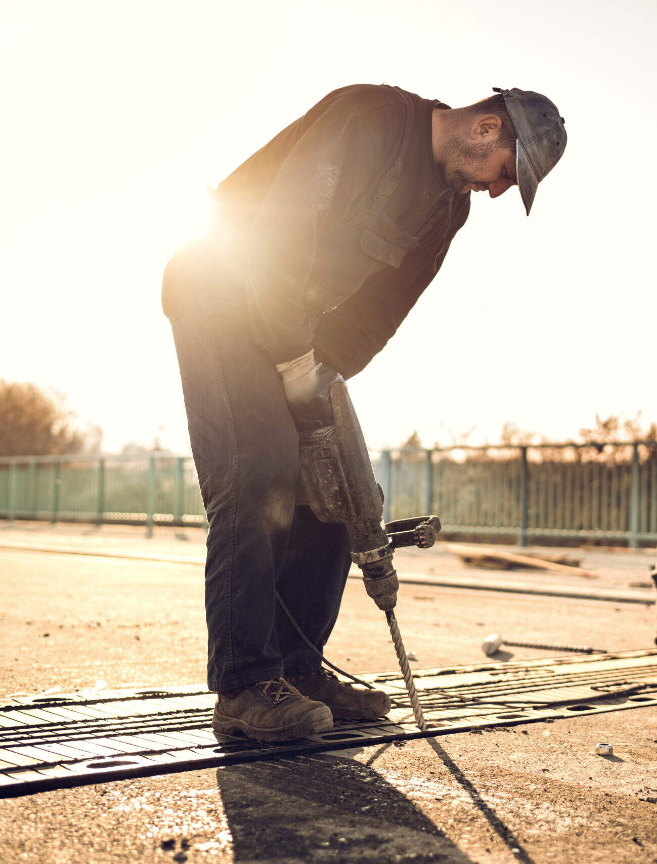 Two manual workers repairing road construction on the bridge at sunset. Focus is on construction worker using a drill.